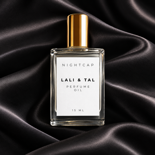 A clear rectangular perfume oil bottle with a white label reading 'NIGHTCAP LALI & TAL PERFUME OIL 15 ML' and a metallic gold cap, centered on draped black satin fabric.