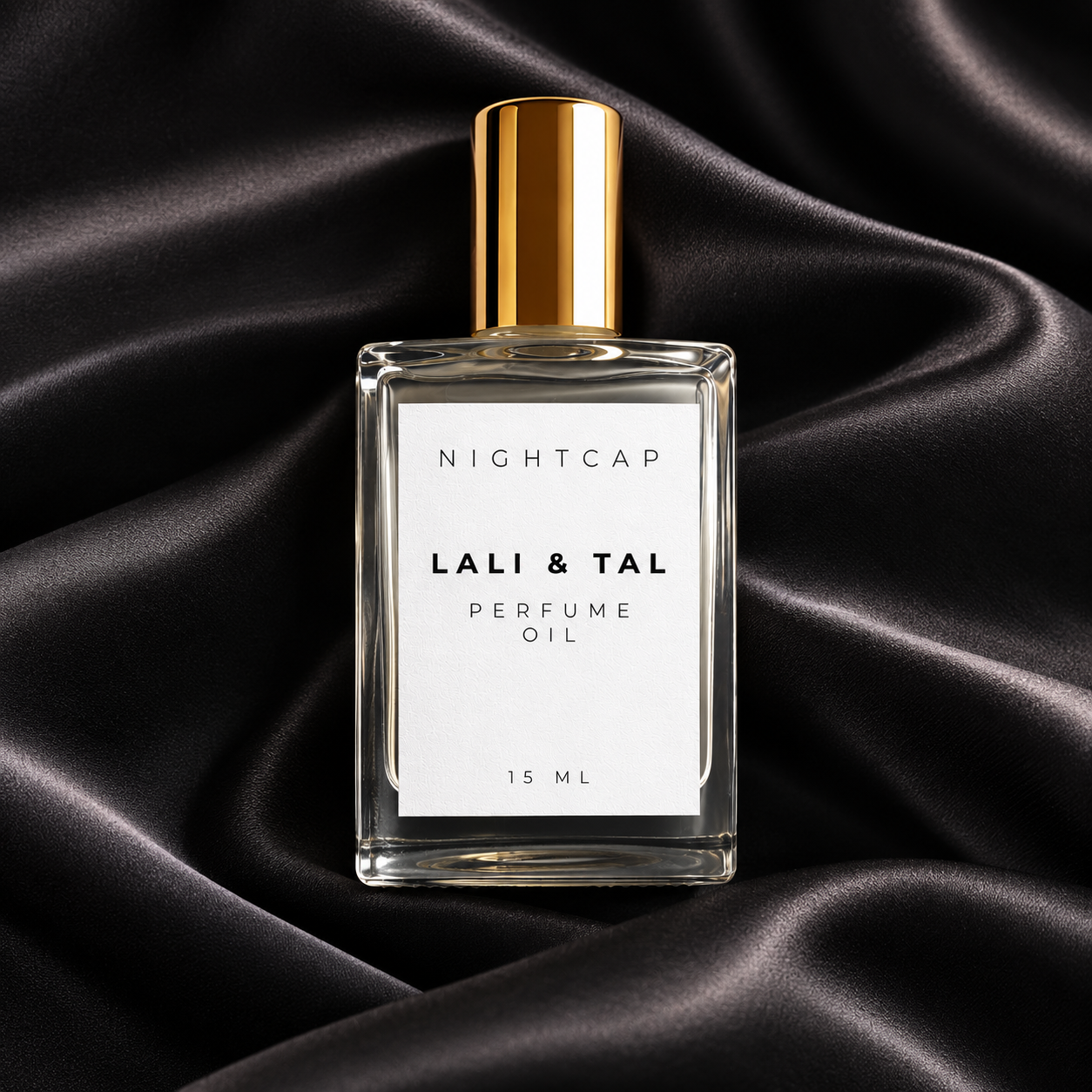 A clear rectangular perfume oil bottle with a white label reading 'NIGHTCAP LALI & TAL PERFUME OIL 15 ML' and a metallic gold cap, centered on draped black satin fabric.