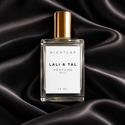 A clear rectangular perfume oil bottle with a white label reading 'NIGHTCAP LALI & TAL PERFUME OIL 15 ML' and a metallic gold cap, centered on draped black satin fabric.