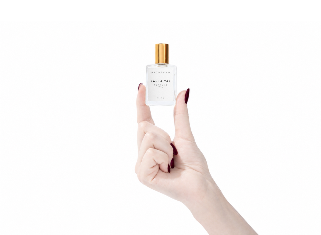 A hand with dark red manicured nails holds up a small clear glass bottle with a gold cap and white label against a solid white background.
